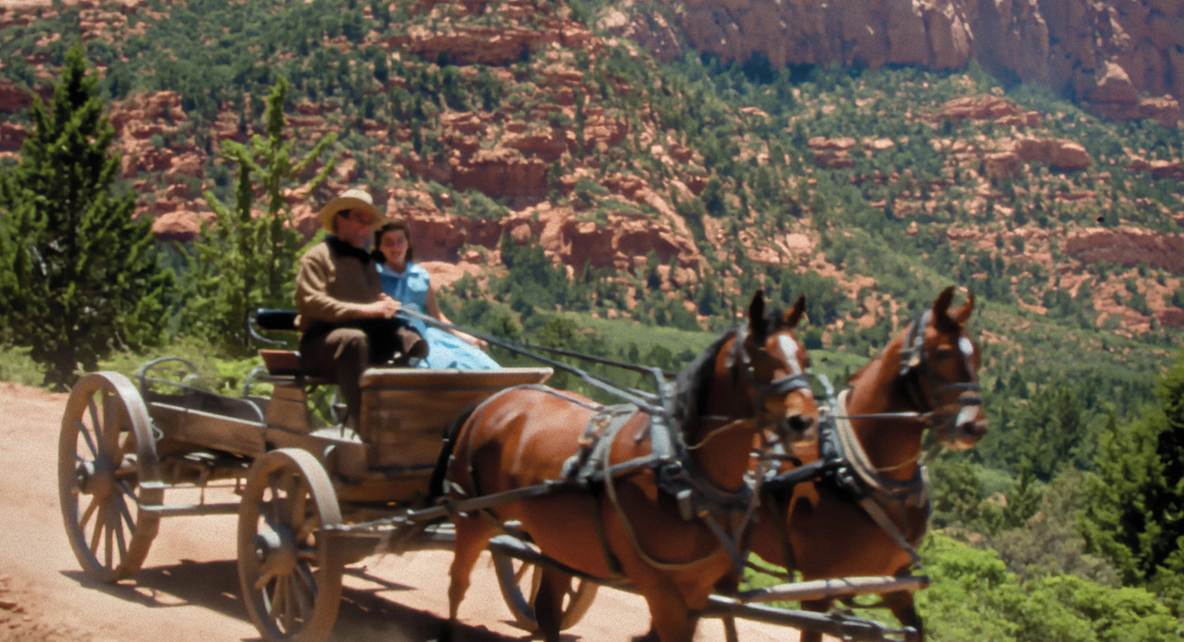 Colourised film still of a horse-drawn carriage crossing a sunlit red-rock landscape