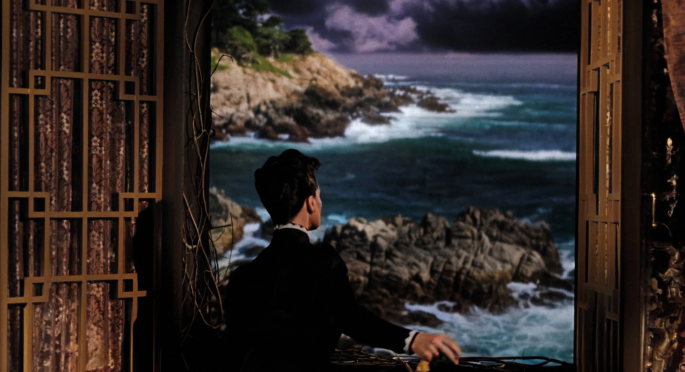Colourised film still of a figure looking through an open window towards a dramatic coastline