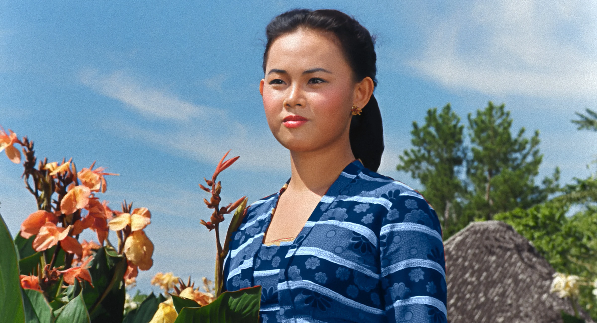 Colourised film still of a woman in blue beside orange flowers in natural daylight